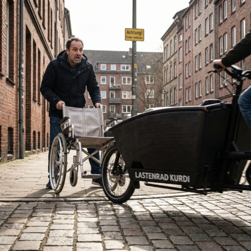 Ein Vater reißt den Rollstuhl seines Sohnes reflexartig zurück, als ein E-Lastenfahrrad haarscharf auf dem Bürgersteig vorbeirast.