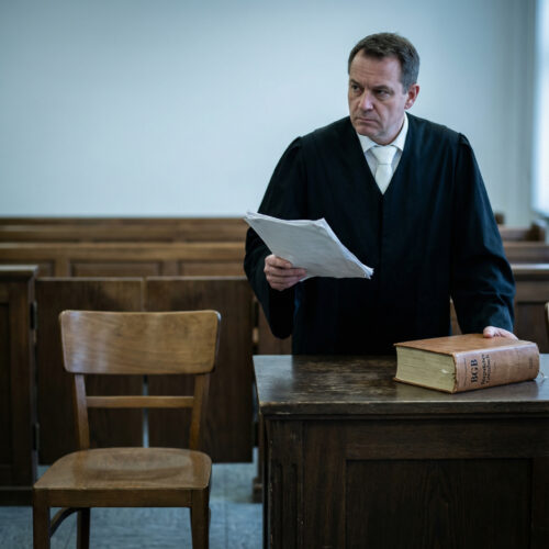 Ein Richter in Robe hält einen dünnen Stapel Dokumente und fixiert den leeren Angeklagtenstuhl im Gerichtssaal.